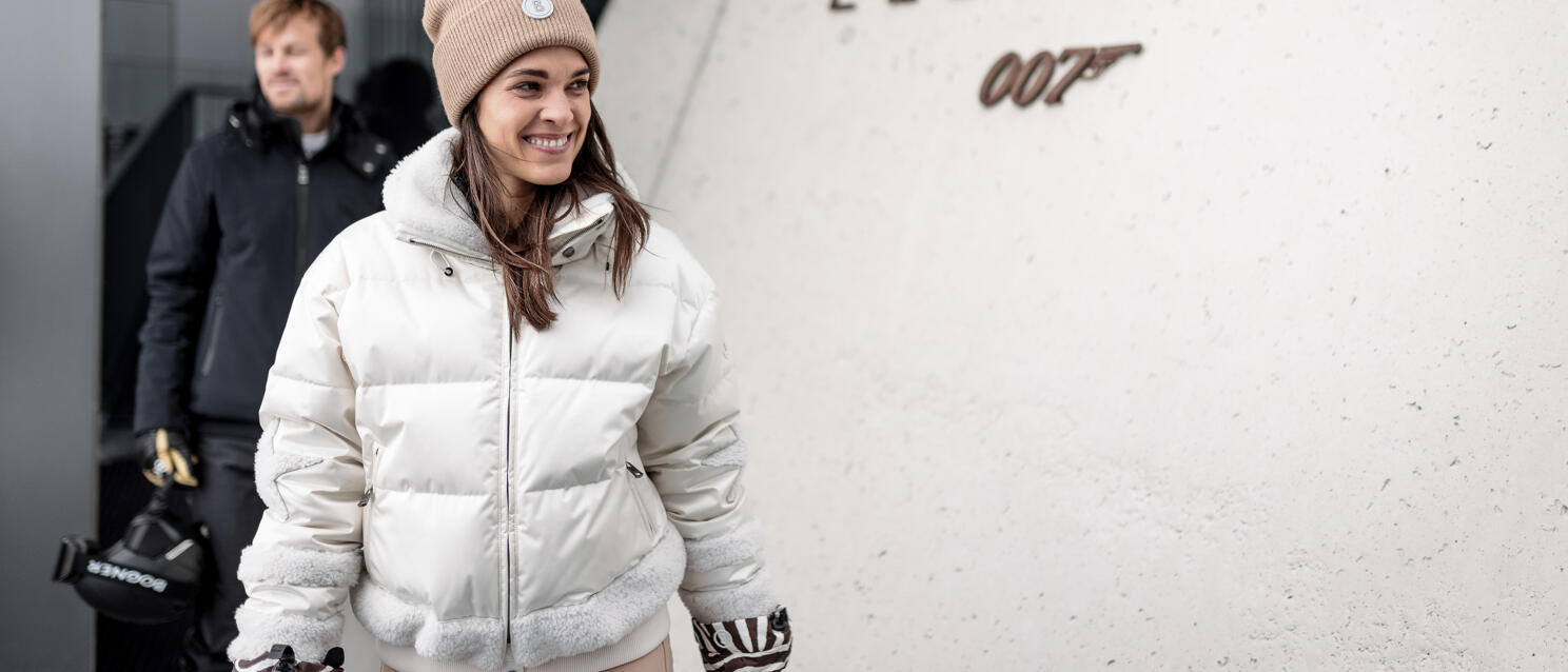 Smiling woman in white down jacket in front of "Elements 007" sign on concrete wall.