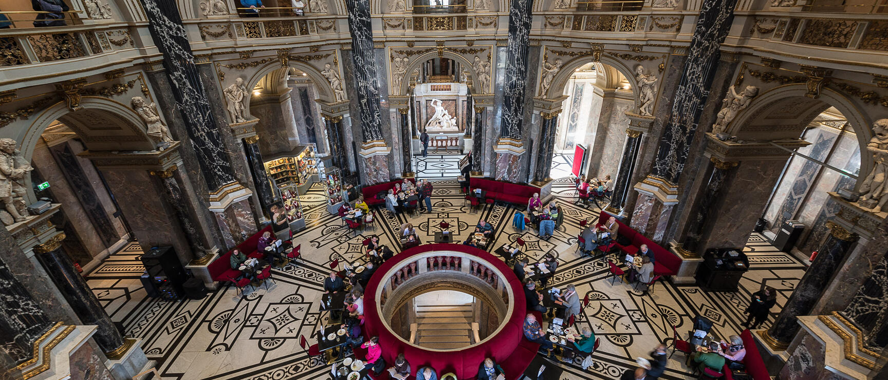 Café in the domed hall of Kunsthistorisches Museum Vienna, red seating benches, marble floor, stucco reliefs, visitors.