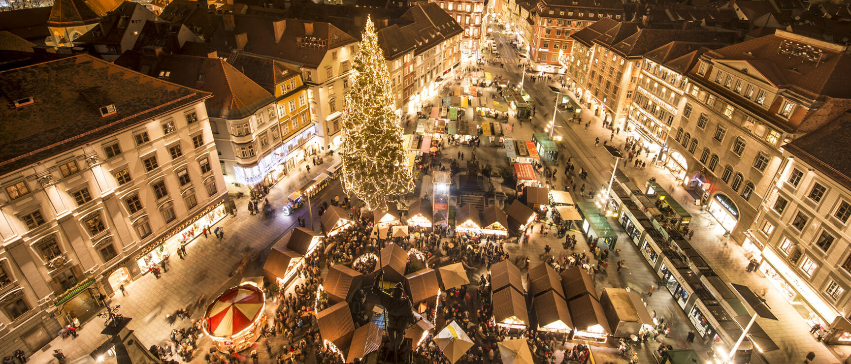 Graz Christkindlmarkt at Hauptplatz at night, large Christmas tree, Schlossberg with clock tower in background.