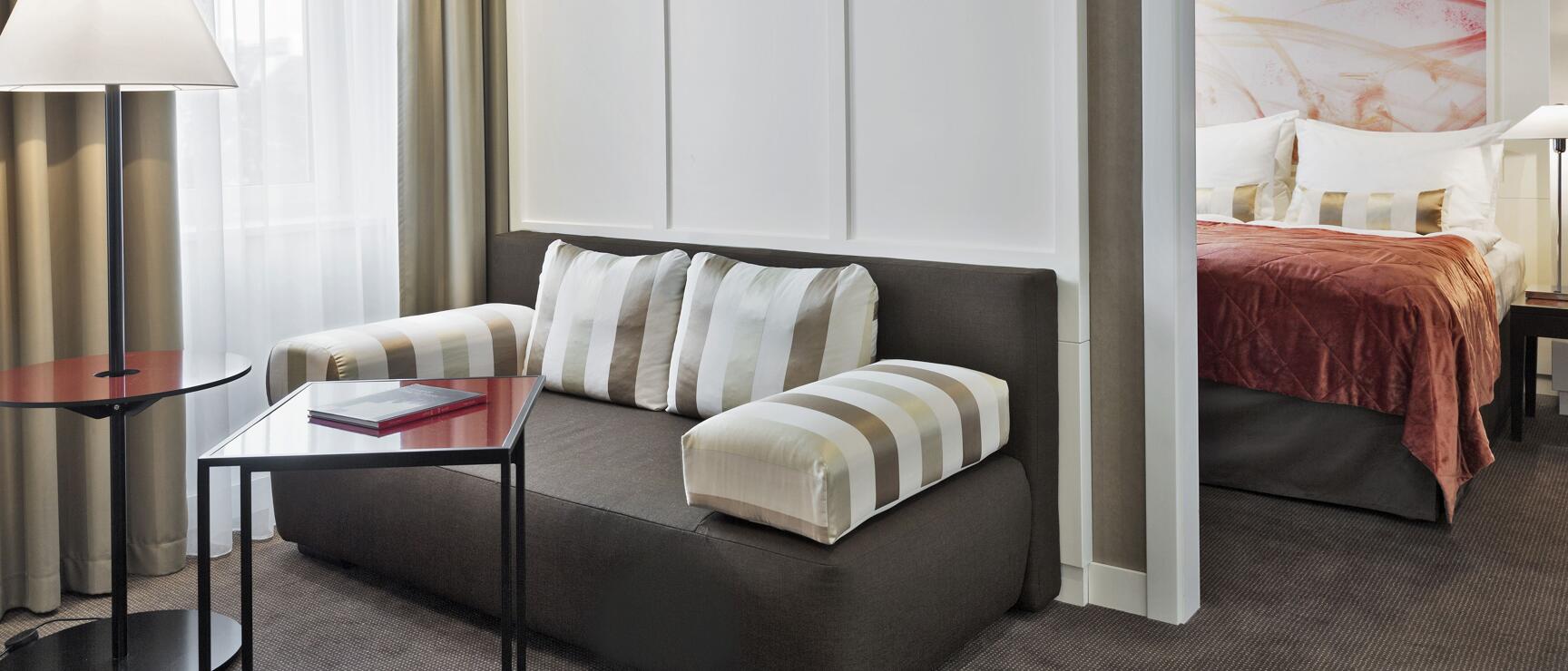 Hotel room with grey sofa, striped cushions and view into bedroom with red bedspread.