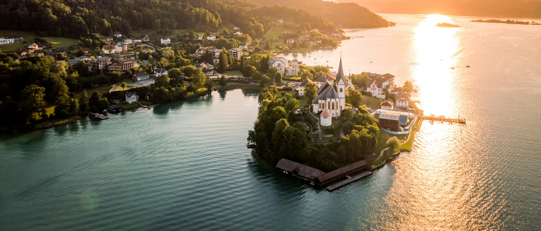 Aerial view of peninsula with white church at Lake Wörthersee at sunset, forested hills in background