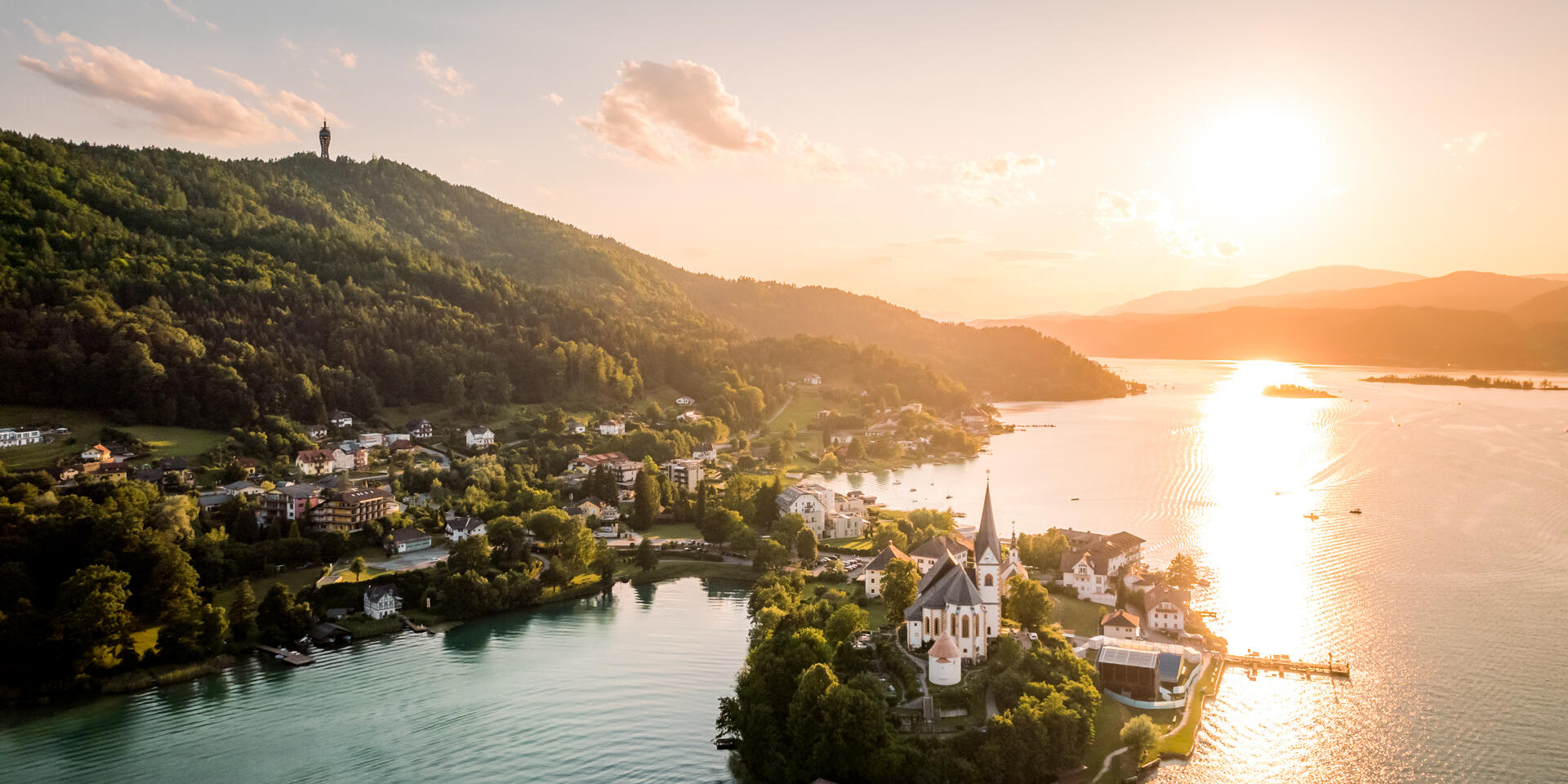 Aerial view of peninsula with white church at Lake Wörthersee at sunset, forested hills in background