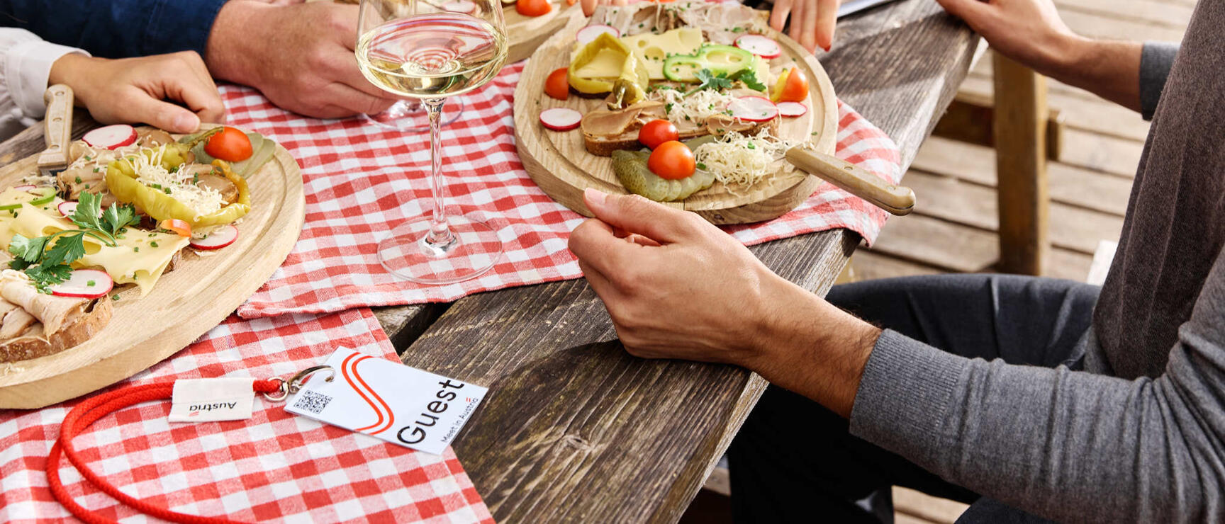 Holztisch mit Brotzeit-Bretter, Weißweinglas und rot-weißer Tischdecke, Hände greifen nach Speisen.