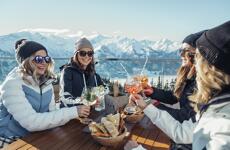 Four women toasting with cocktails on a sunny terrace with snow-capped mountains in background.