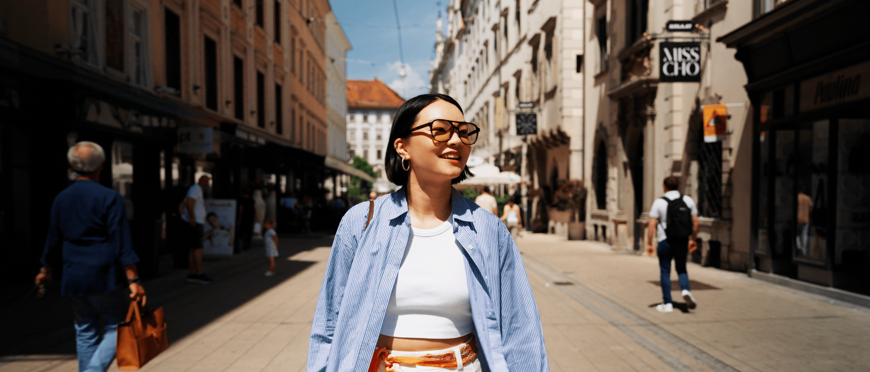 Young woman wearing sunglasses in a sunny pedestrian street with shops and passersby in the background.