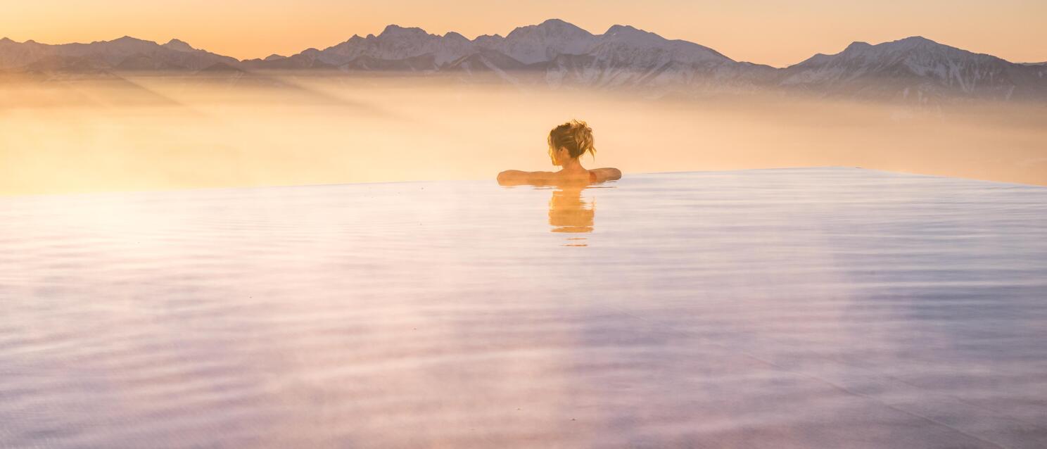 Woman in infinity pool at sunset, snow-capped mountain range and morning mist in background.