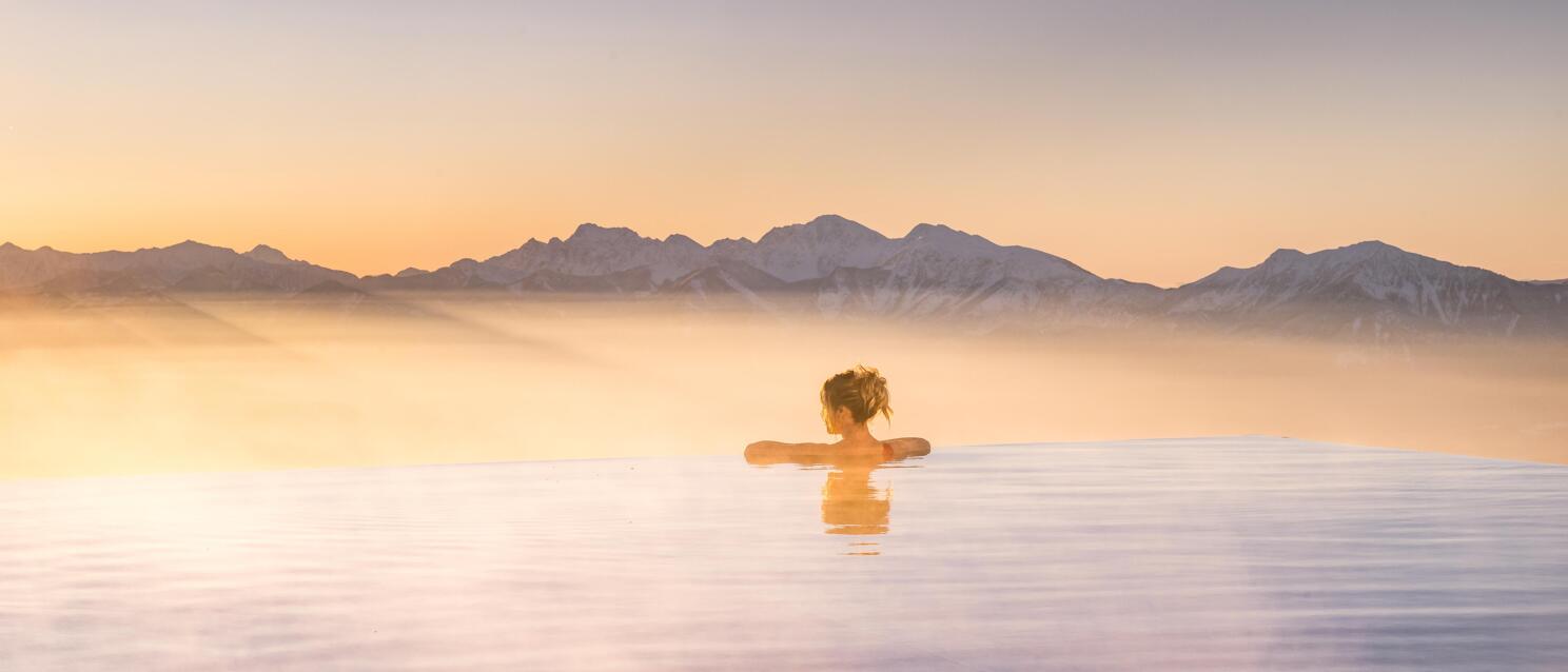 Woman in infinity pool at sunset, snow-capped mountain range and morning mist in background.