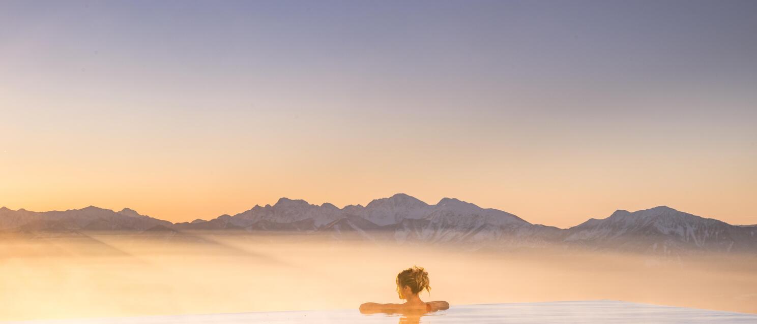 Woman in infinity pool at sunset, snow-capped mountain range and morning mist in background.