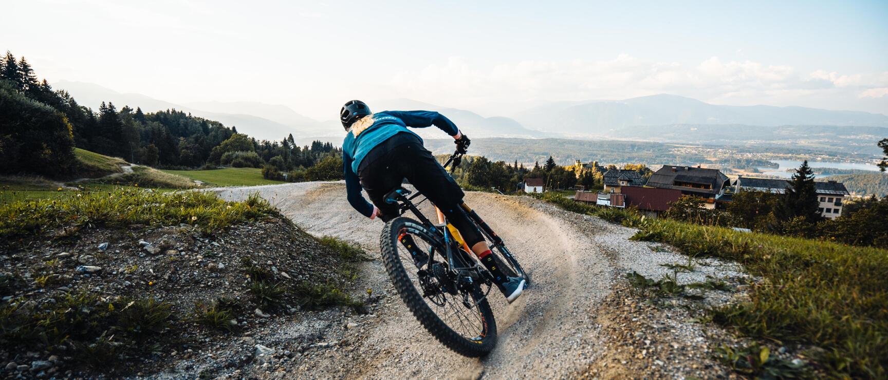 Mountain biker cornering on gravel trail with valley, lake and mountain range in the background.