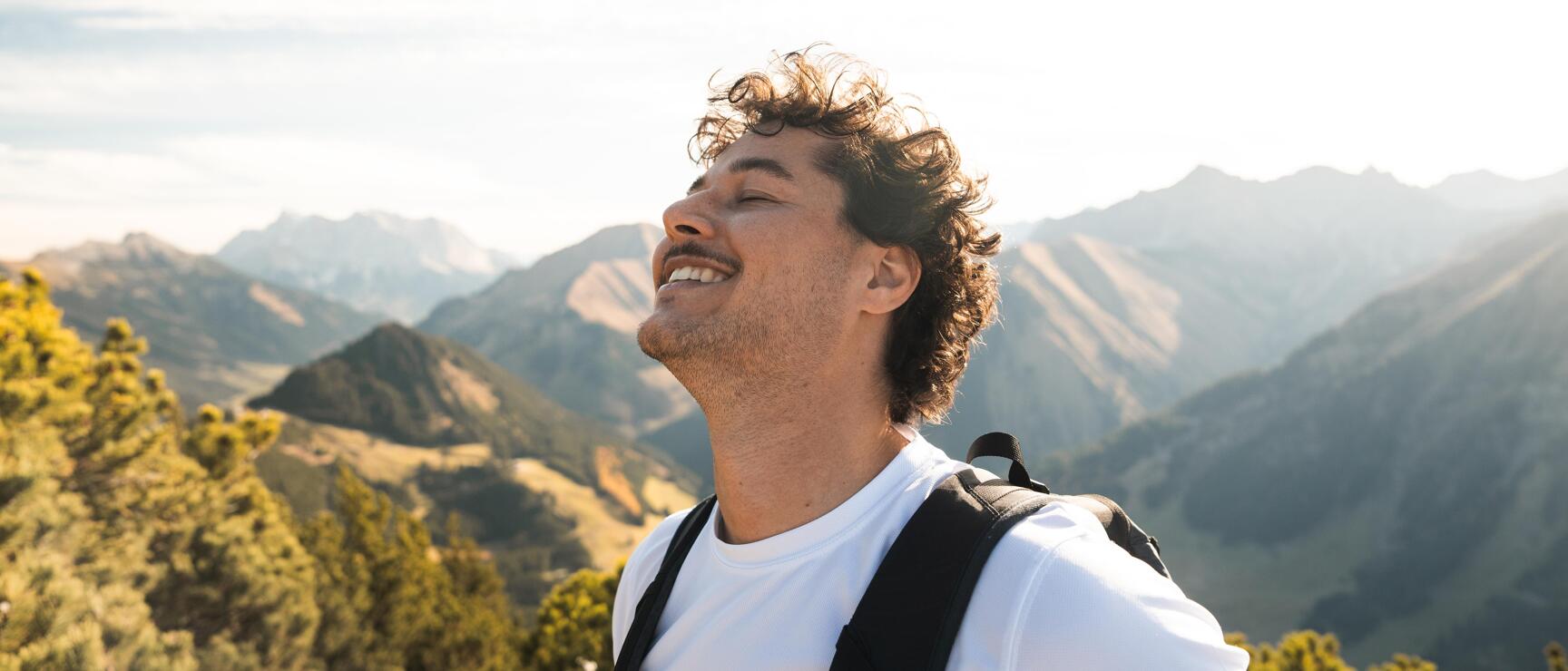 Man with backpack and white t-shirt on a mountain peak, alpine panorama in the background.
