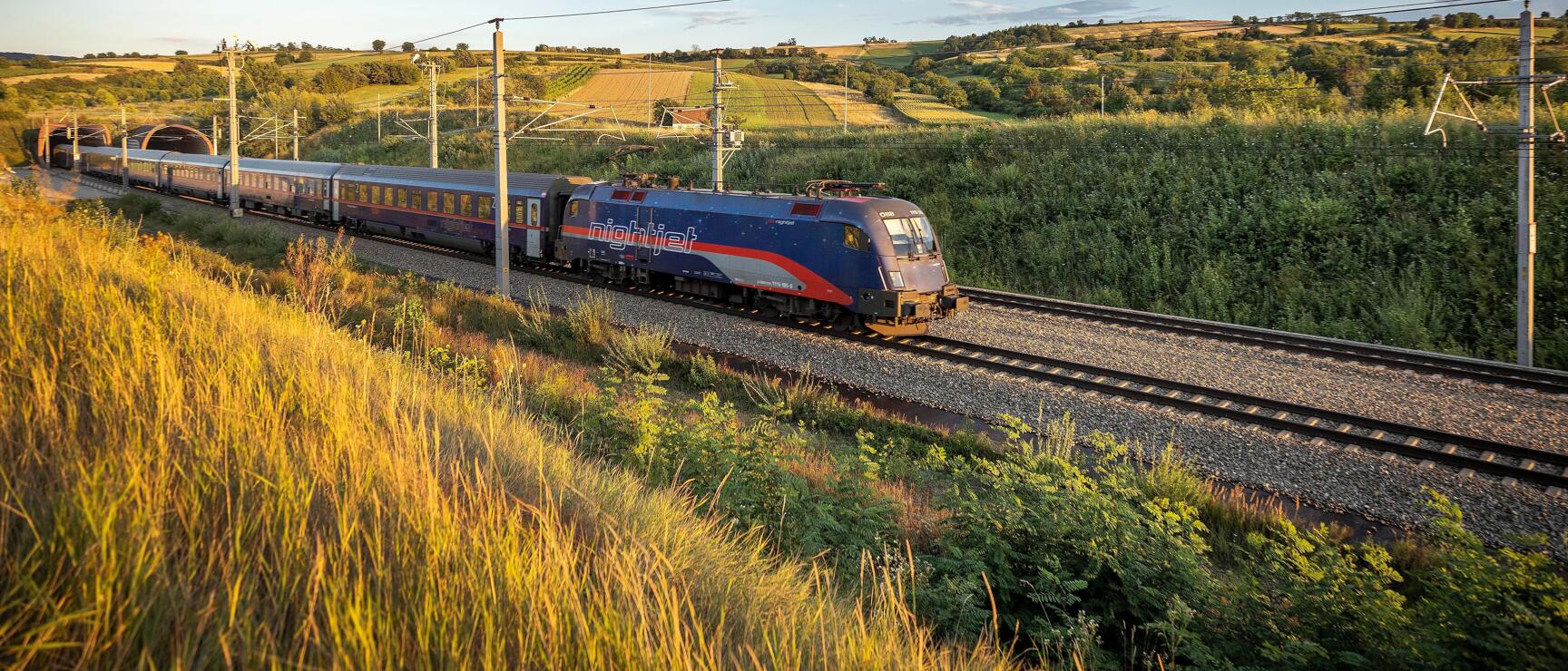 Nightjet train traveling through hilly landscape with fields and meadows at evening light