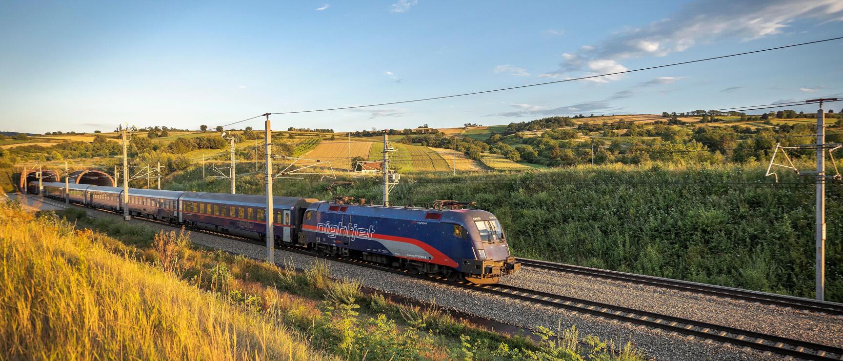 Nightjet train traveling through hilly landscape with fields and meadows at evening light