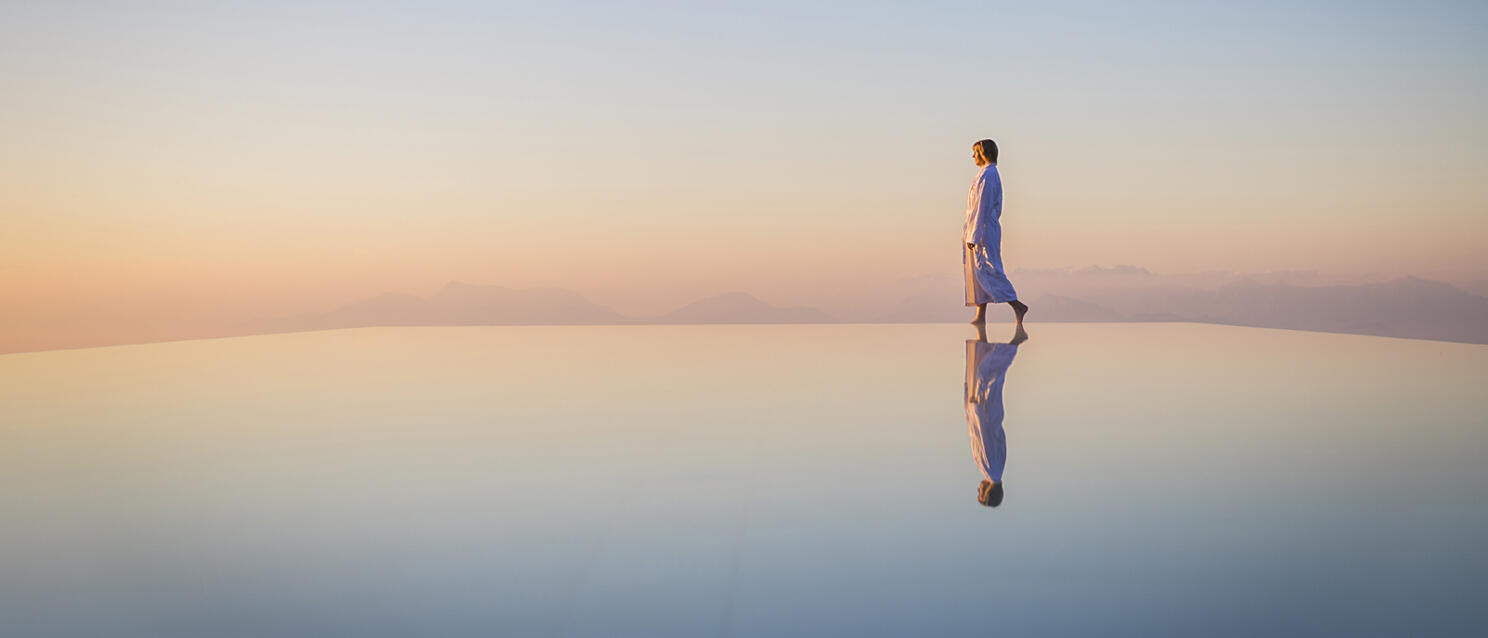 Person in white bathrobe walking at the edge of an infinity pool, water reflection, pastel-coloured evening sky.