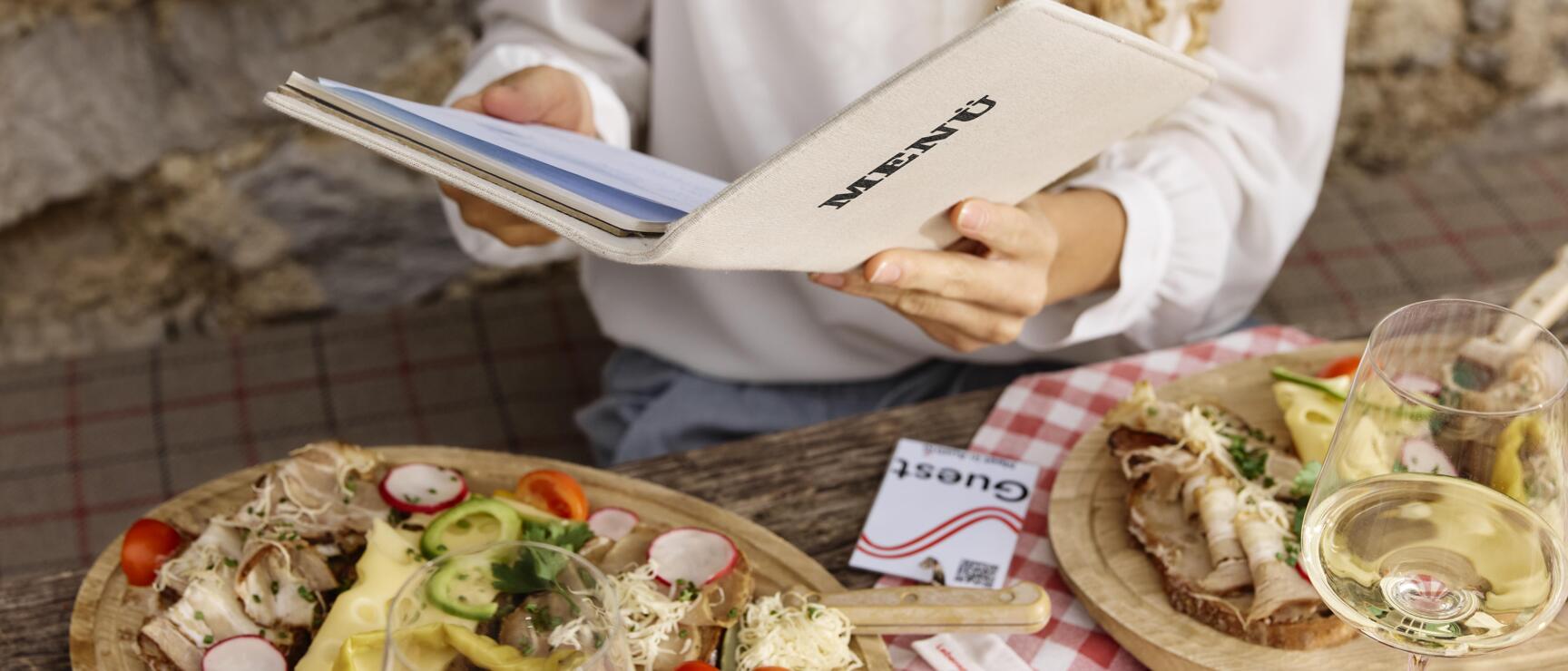 Woman holding menu card with wooden boards of bread, vegetables and white wine on checkered tablecloth