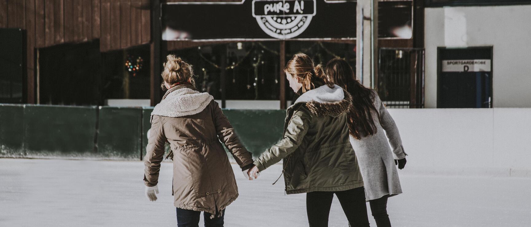 Three women in winter jackets running hand in hand on ice rink, building with 'pure A!' sign in background.