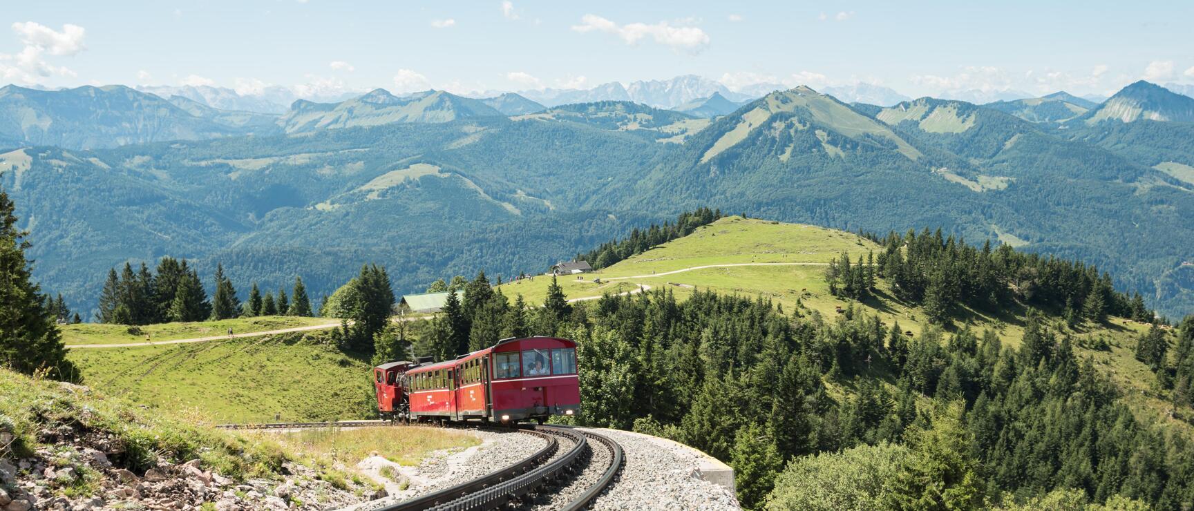 Red cogwheel train on winding mountain route, green Alpine landscape and mountain panorama in background.
