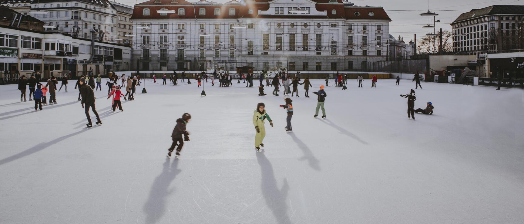 Many people and children ice skating on an outdoor ice rink in front of a white city building in Vienna.