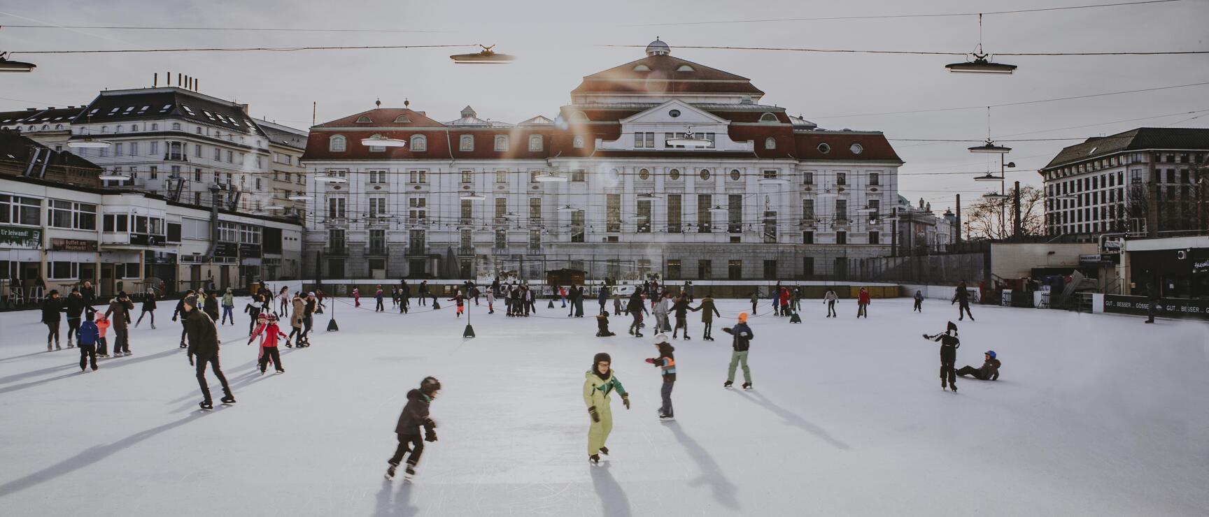 Many people and children ice skating on an outdoor ice rink in front of a white city building in Vienna.