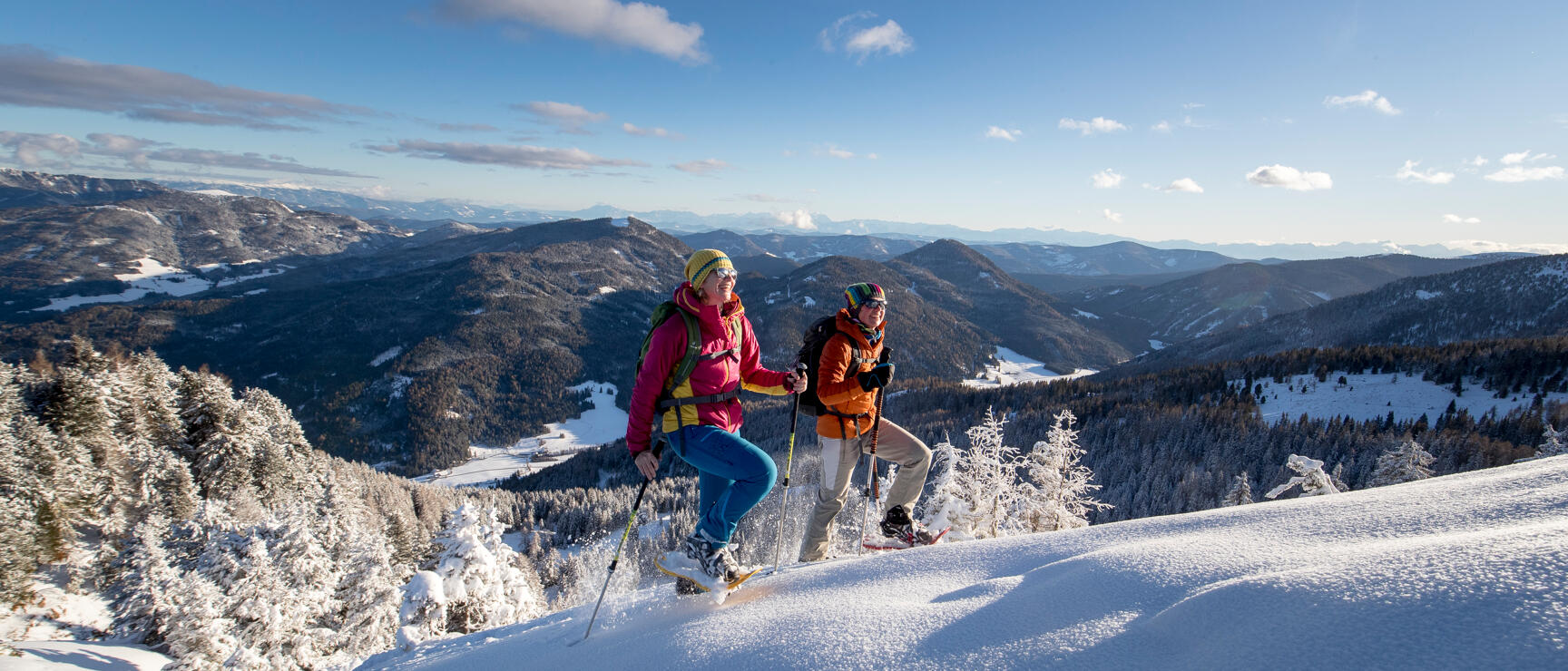 Two people with snowshoes on snowy slope, mountain panorama and forests in background.