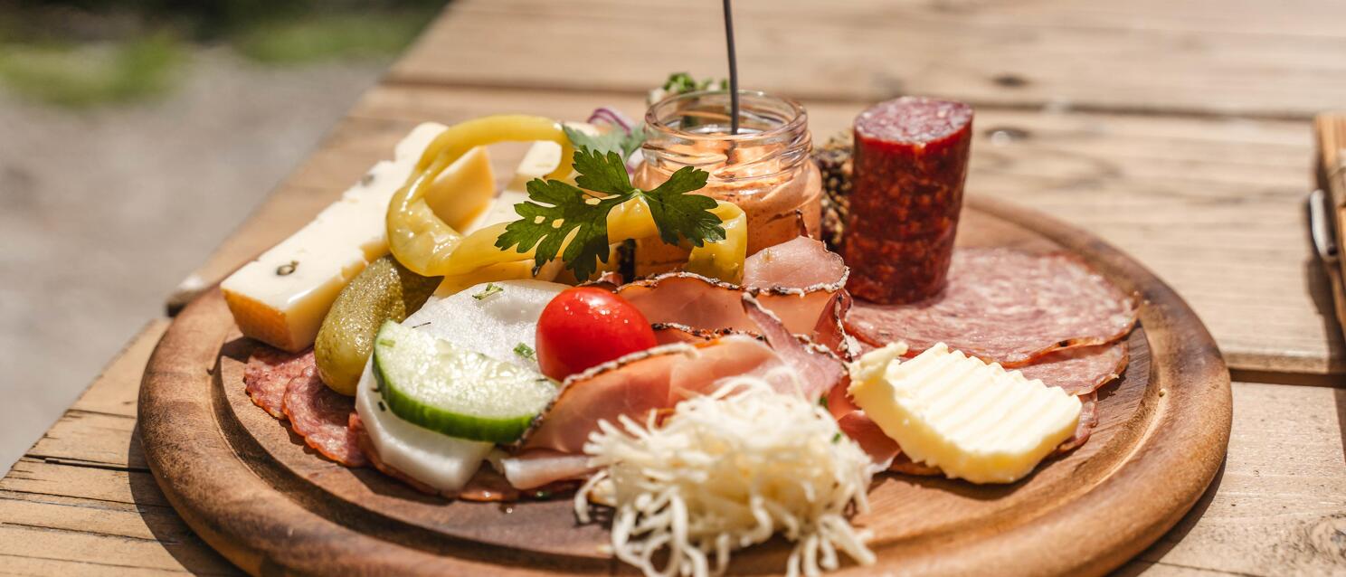 Brettljause on round wooden board: ham, salami, cheese, butter, cucumber, tomato, peppers and mustard in jar.