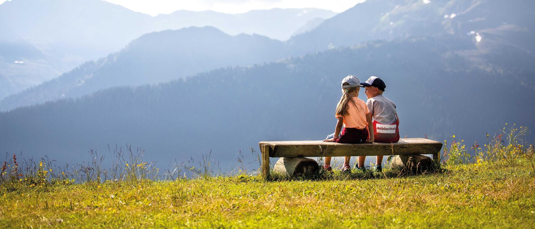 Two children sitting on a wooden bench at the Wiedersberger Horn in Alpbach, looking out over a flowering mountain meadow and the surrounding Tyrolean mountain landscape