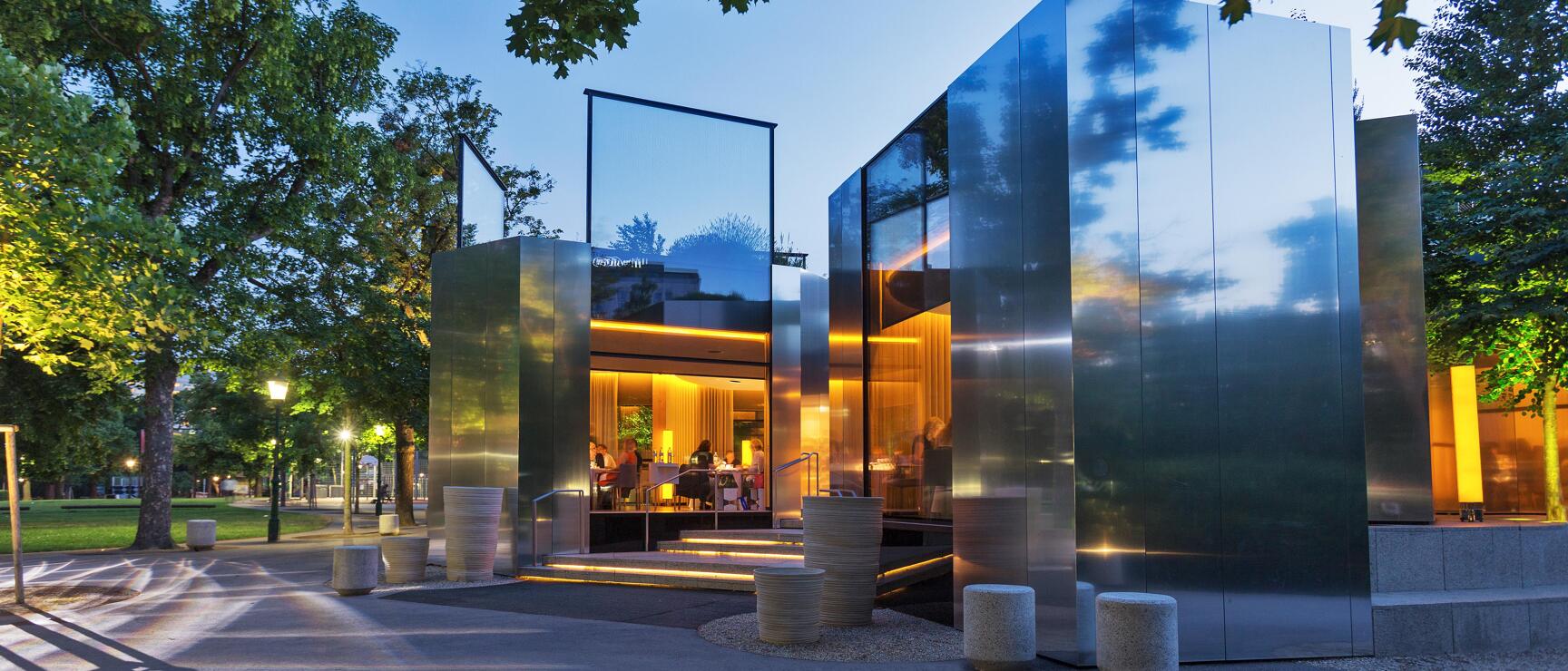 Modern glass pavilion with mirror façade in park at dusk, warm lighting inside, people visible.