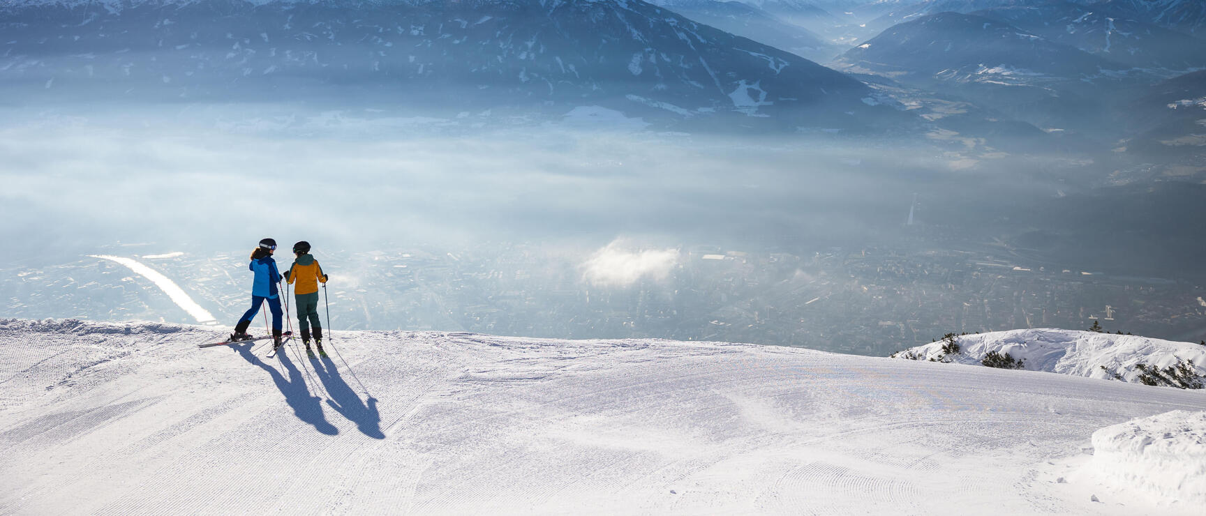 Two skiers on snowy slope with view of Innsbruck in valley, Alpine panorama in background.