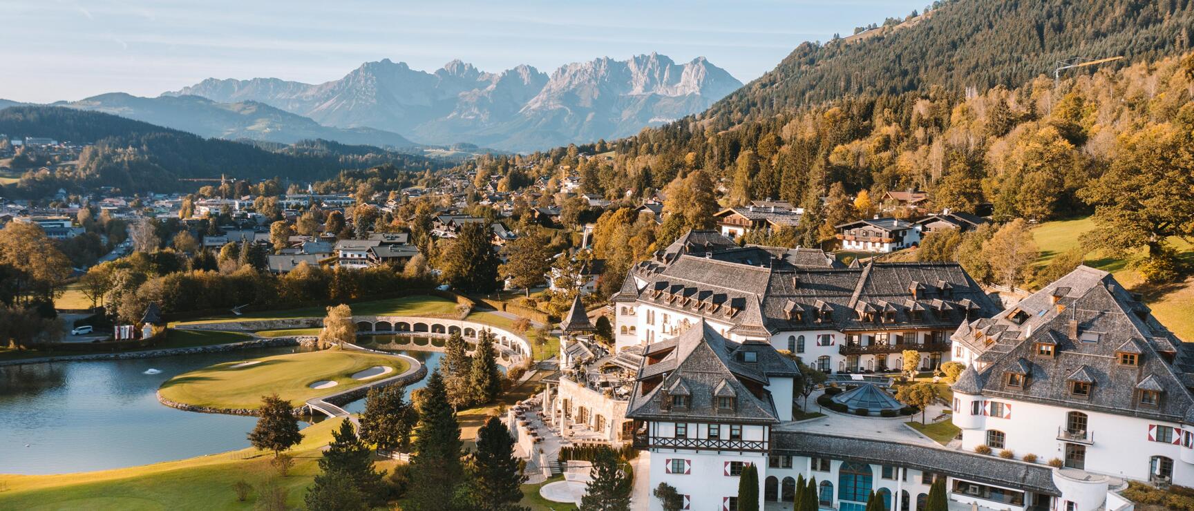 Aerial view of a white hotel complex with pool and golf course, lake and mountain panorama in autumn.