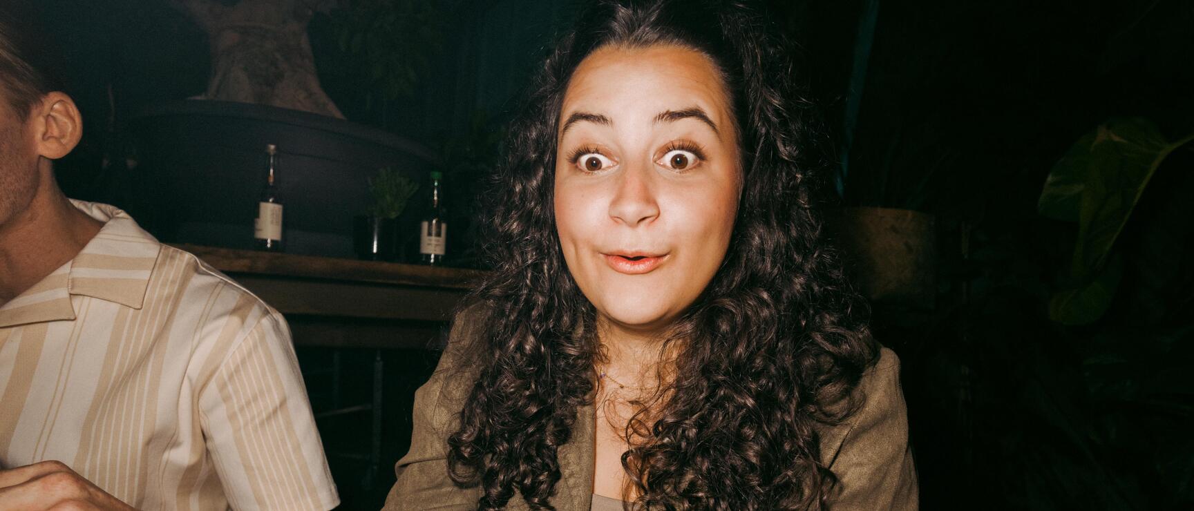 Frau mit lockigem Haar und aufgerissenem Blick sitzt in einem dunklen Restaurant, vor ihr ein Teller mit Speiseresten.