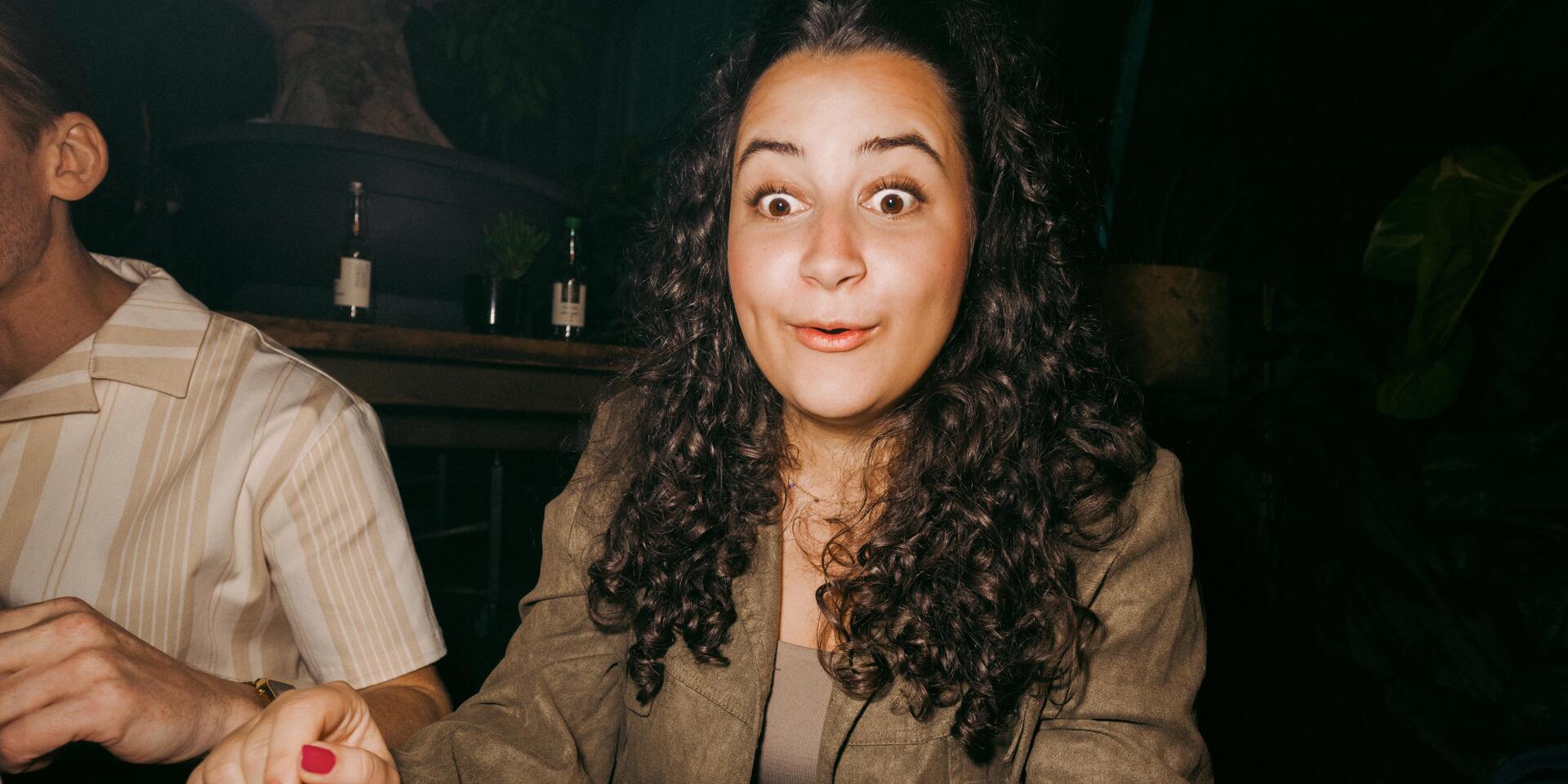 Woman with curly hair and wide-eyed expression sitting in a dark restaurant, with a plate of food scraps in front of her.