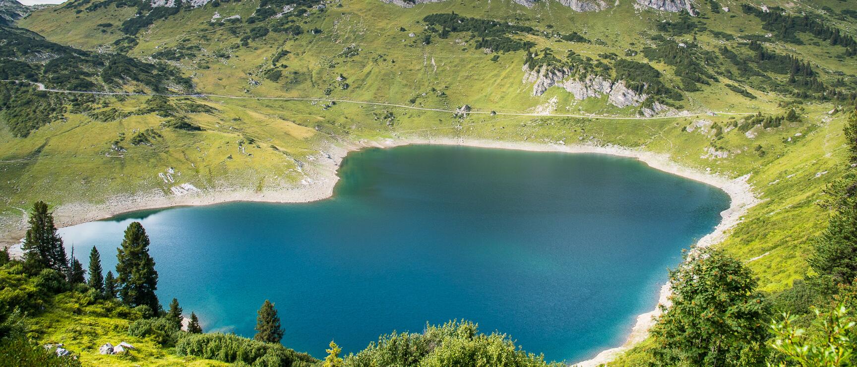 Turquoise mountain lake surrounded by green alpine meadows, rocky peaks in background, blue sky