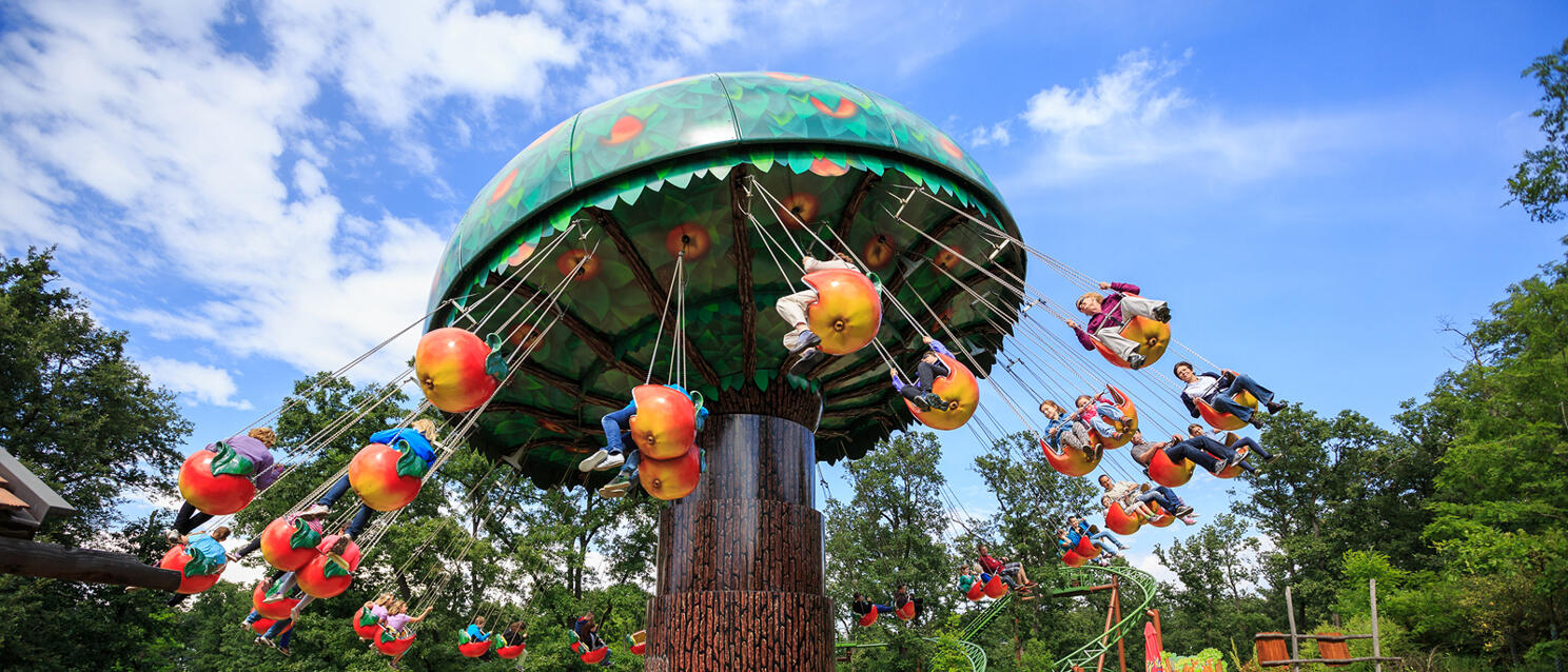 Chain carousel in apple shape at amusement park with visitors watching and trees in background.