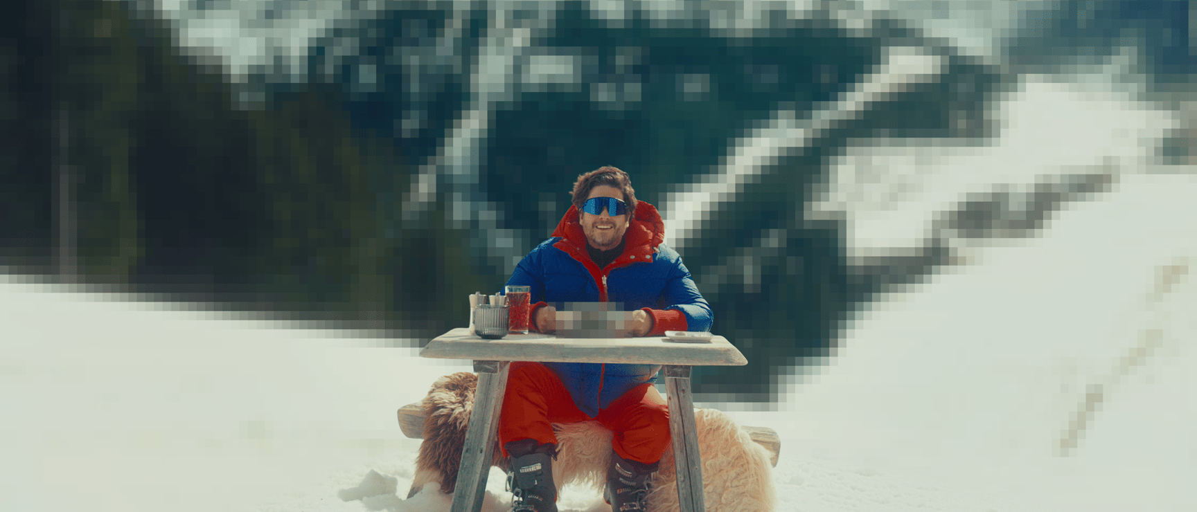 Mann in blau-roter Skijacke sitzt an Holztisch im Schnee, Getränke auf dem Tisch, schneebedeckte Berge im Hintergrund.