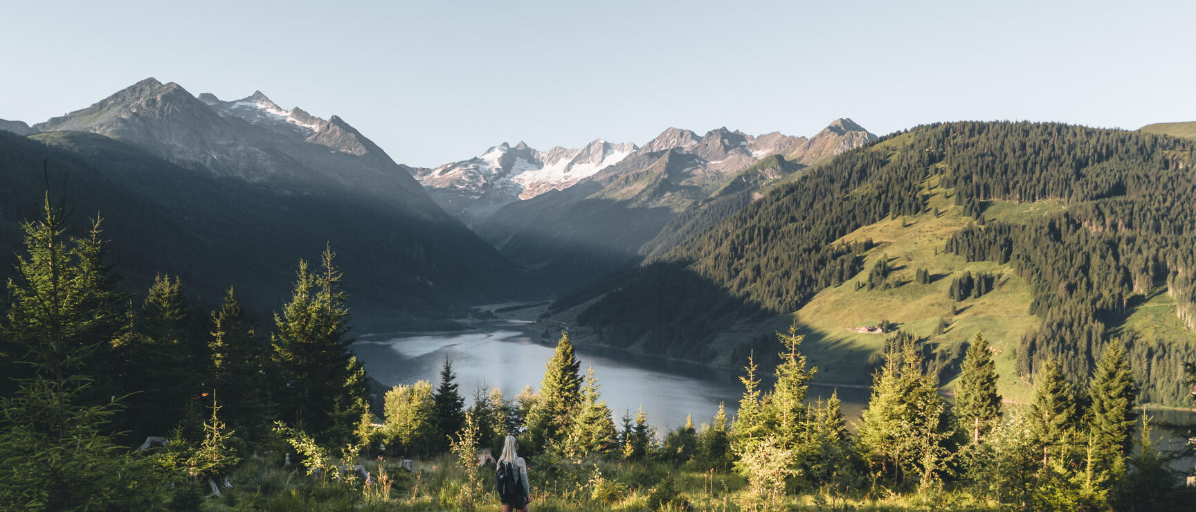 Hiker with backpack standing on hilltop, viewing mountain lake and snow-capped peaks, coniferous forest in foreground.