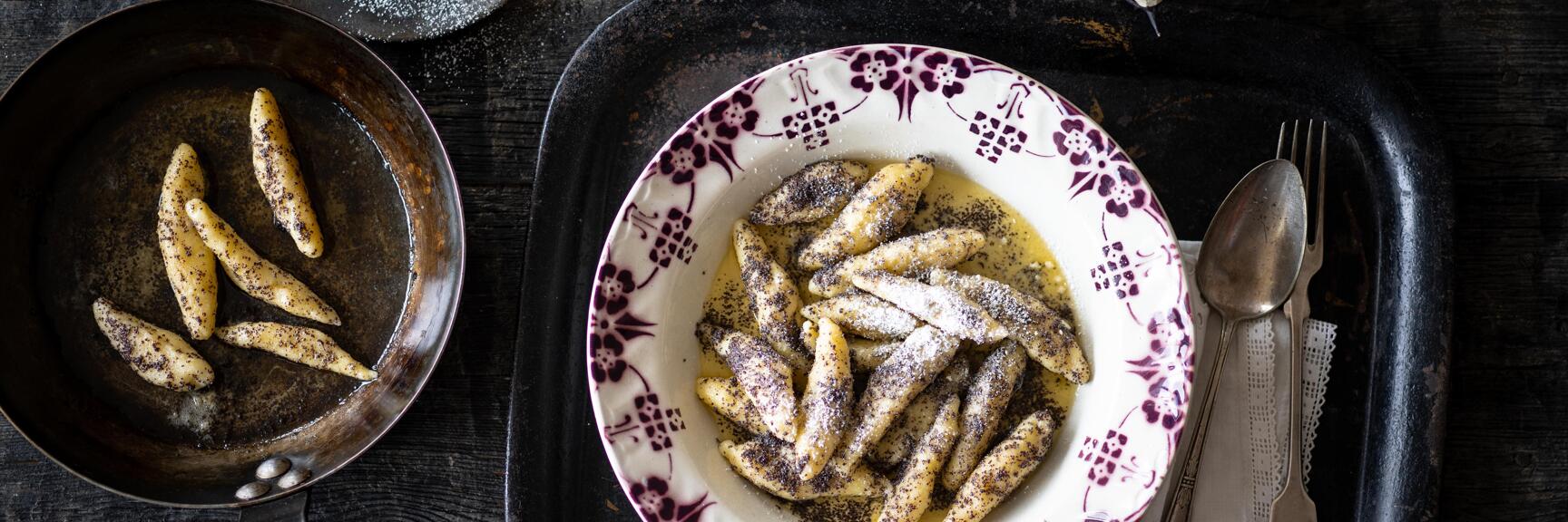 Schupfnudeln with poppy seeds and icing sugar on a floral plate, pan and sugar sifter beside, top view.