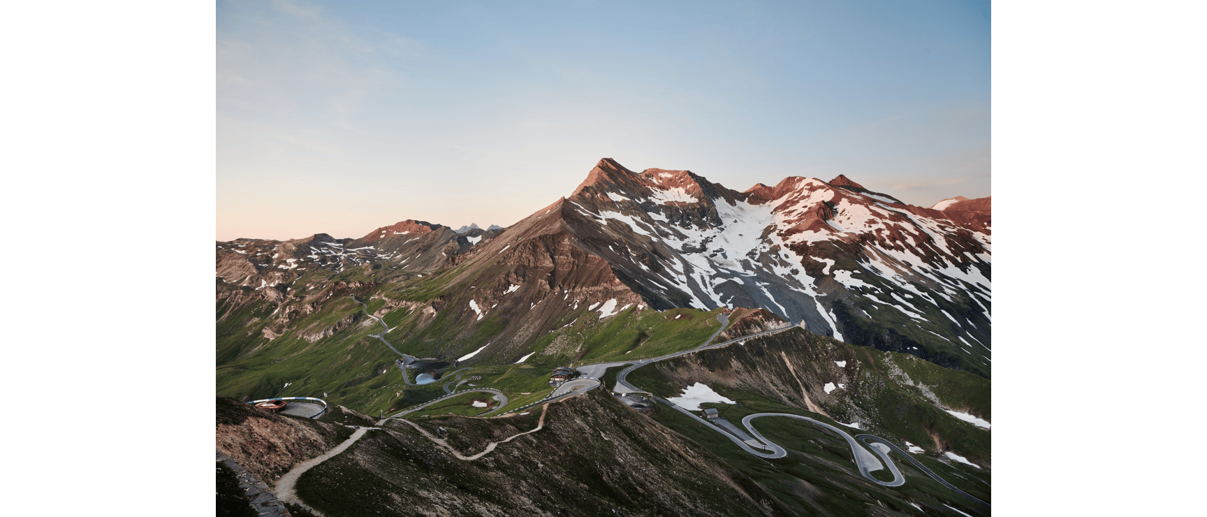 Winding Großglockner High Alpine Road with hairpin turns, mountain hut and snow-capped peaks in sunrise light.