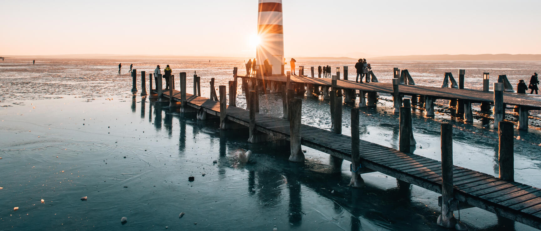 Red and white lighthouse on wooden pier of Lake Neusiedl at sunset, frozen water surface, people on the pier.