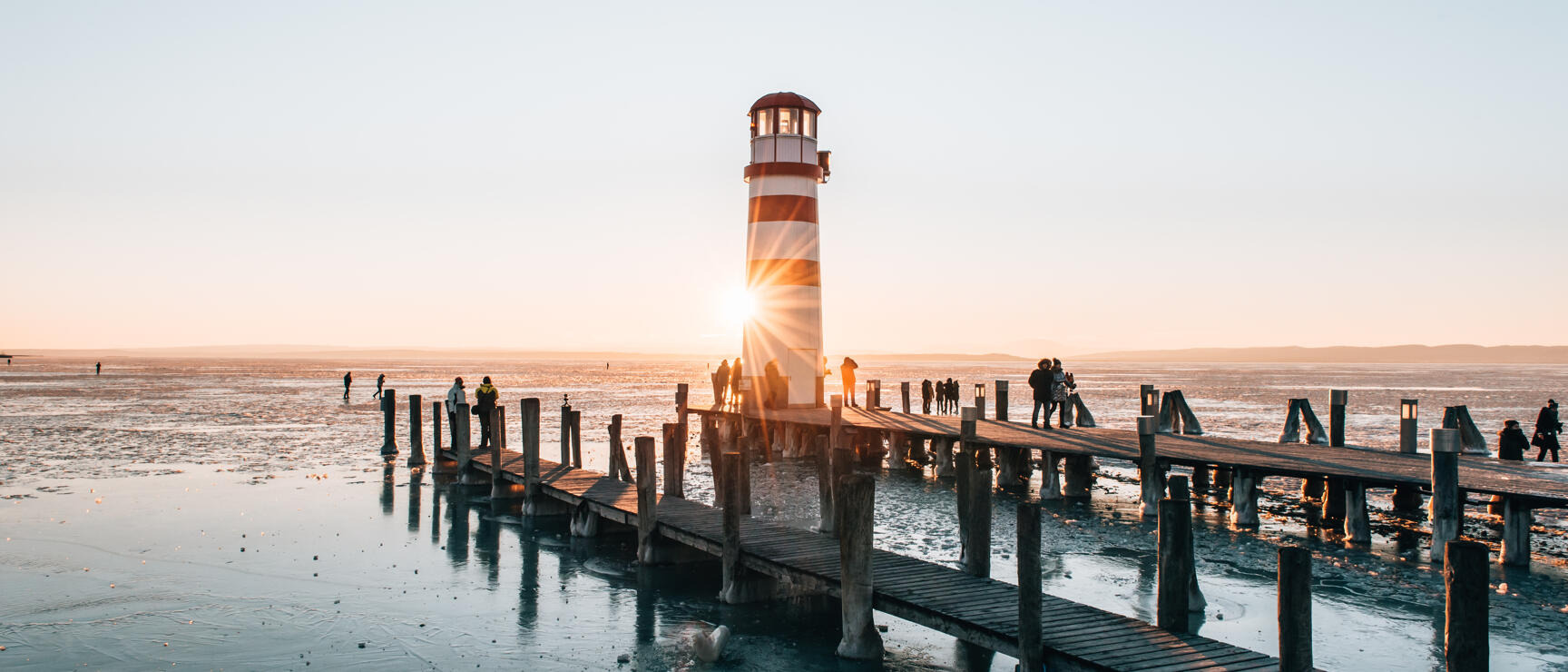 Rot-weißer Leuchtturm am Holzsteg des Neusiedler Sees bei Sonnenuntergang, vereiste Wasserfläche, Personen auf dem Steg.