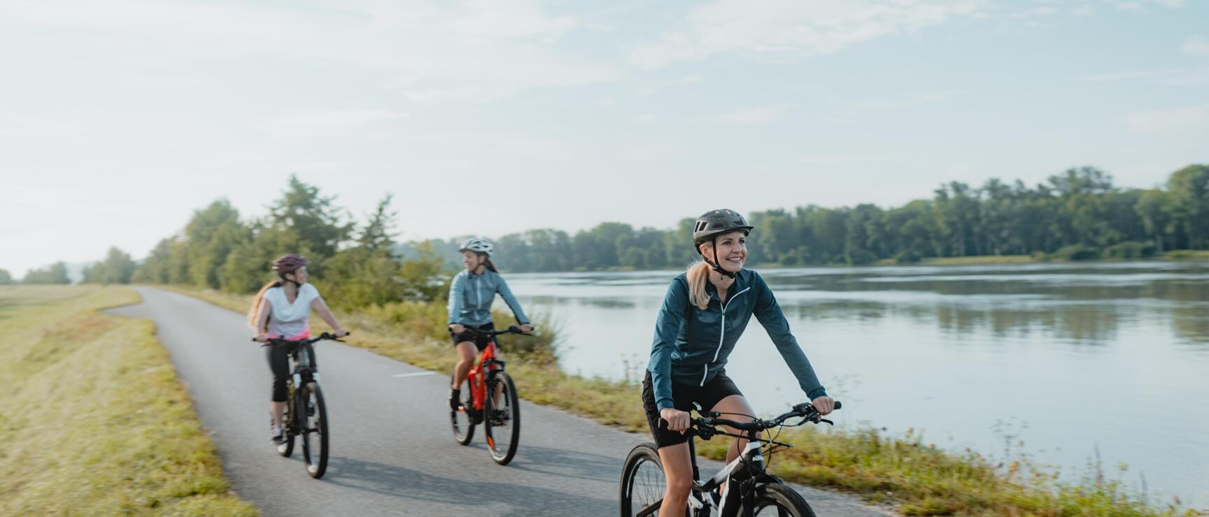Three female cyclists wearing helmets on an asphalt path along a river with trees in the background.
