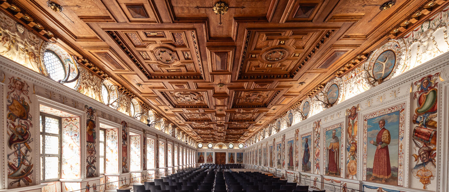 Grand hall with coffered ceiling, frescoes on walls and rows of chairs on marble floor.