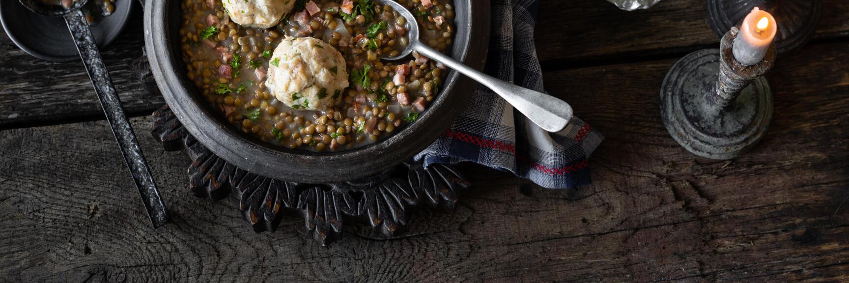 Linsensuppe mit Knödeln in einer Tonschüssel, Rotwein, Kerzen und Petersilie auf rustikalem Holztisch.