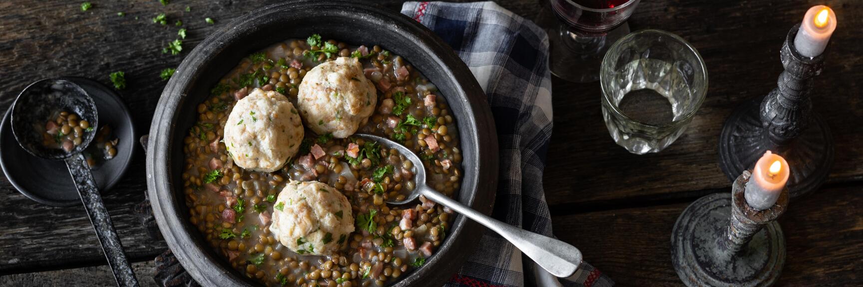 Lentil soup with dumplings in a clay bowl, red wine, candles and parsley on a rustic wooden table.