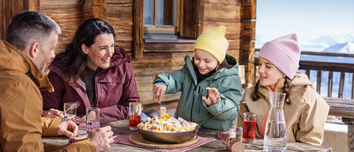 Two adults and two children eating Kaiserschmarrn on an alpine hut terrace, mountains in the background.