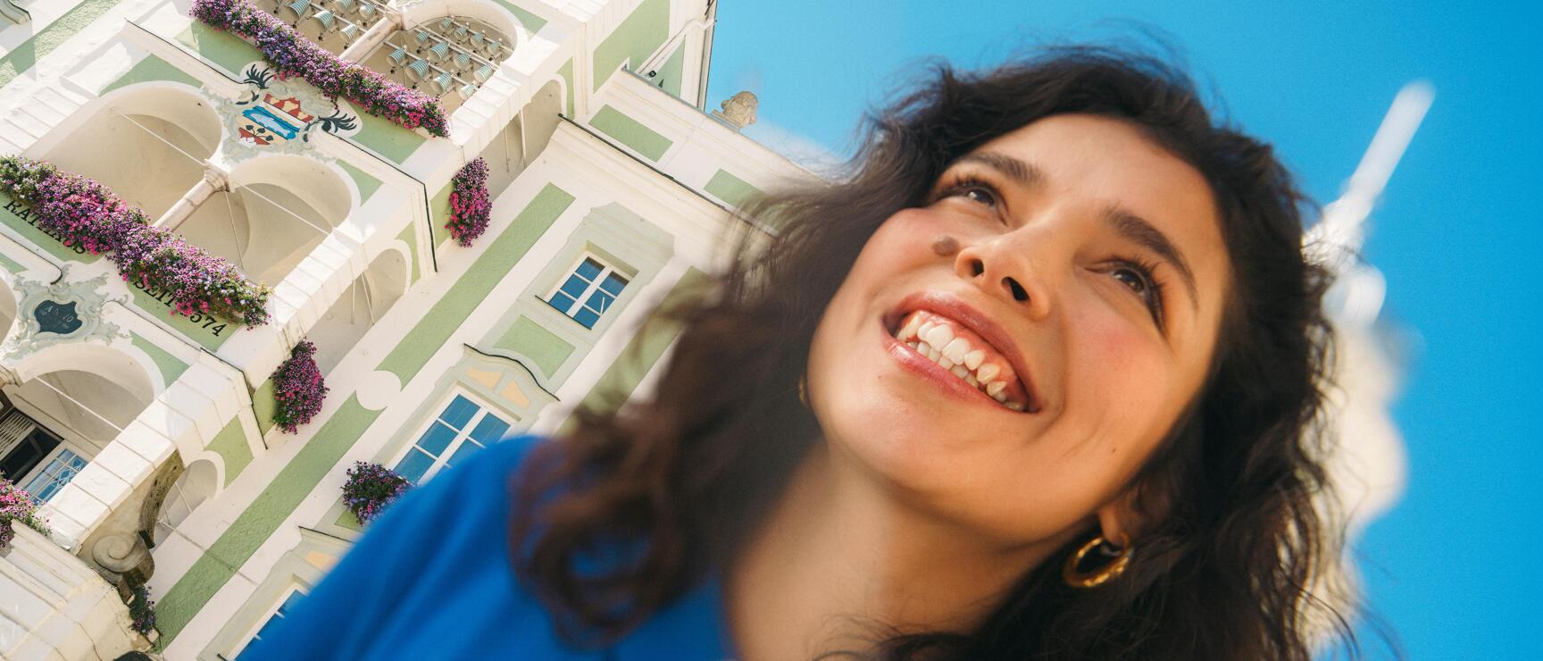 Smiling woman in front of green-white town hall with flower decorations and clock in Gmunden, blue sky.