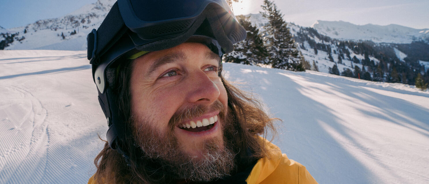 Laughing man with ski helmet and yellow jacket on snowy ski run, snow-covered mountains in background.