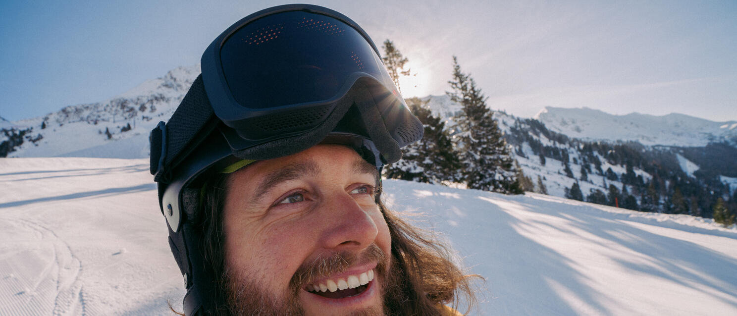 Laughing man with ski helmet and yellow jacket on snowy ski run, snow-covered mountains in background.