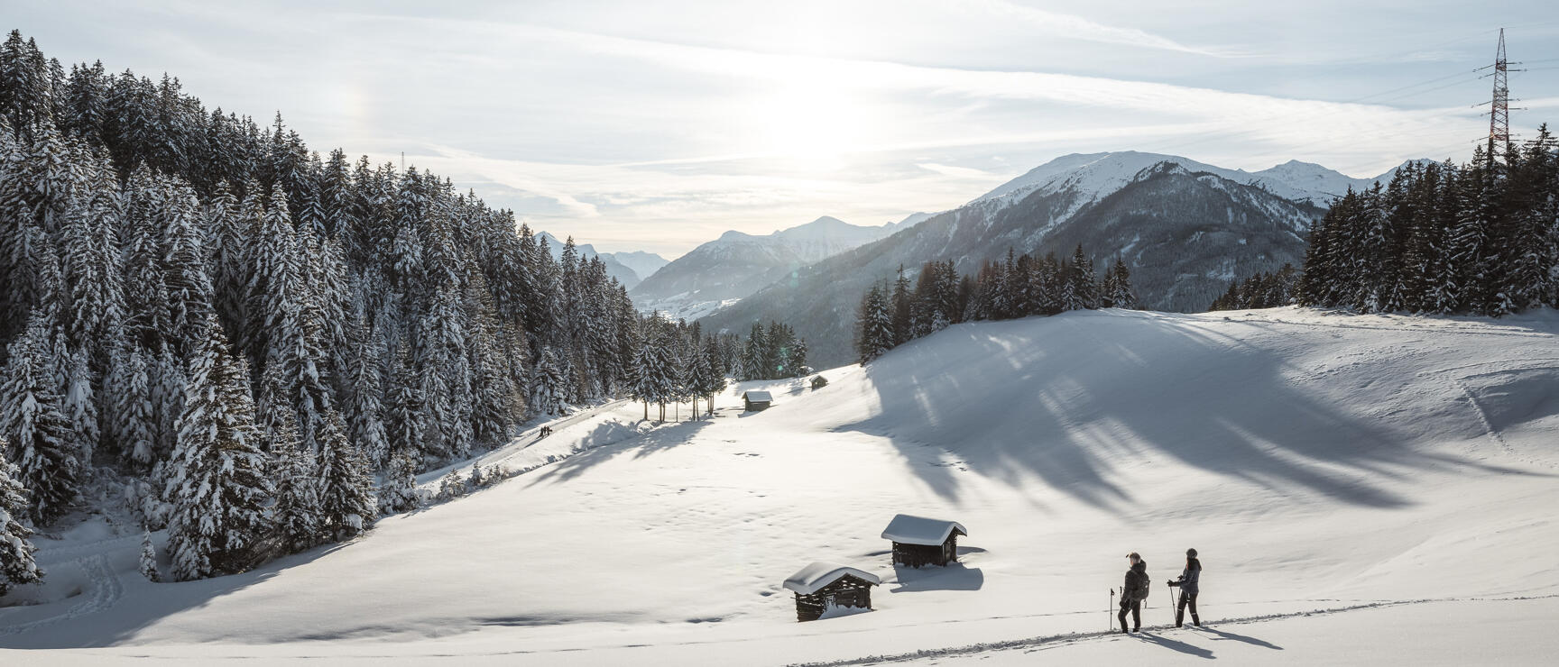 Two people wearing snowshoes on a snowy alpine meadow with small wooden cabins, snowy forest and mountain panorama in the background.