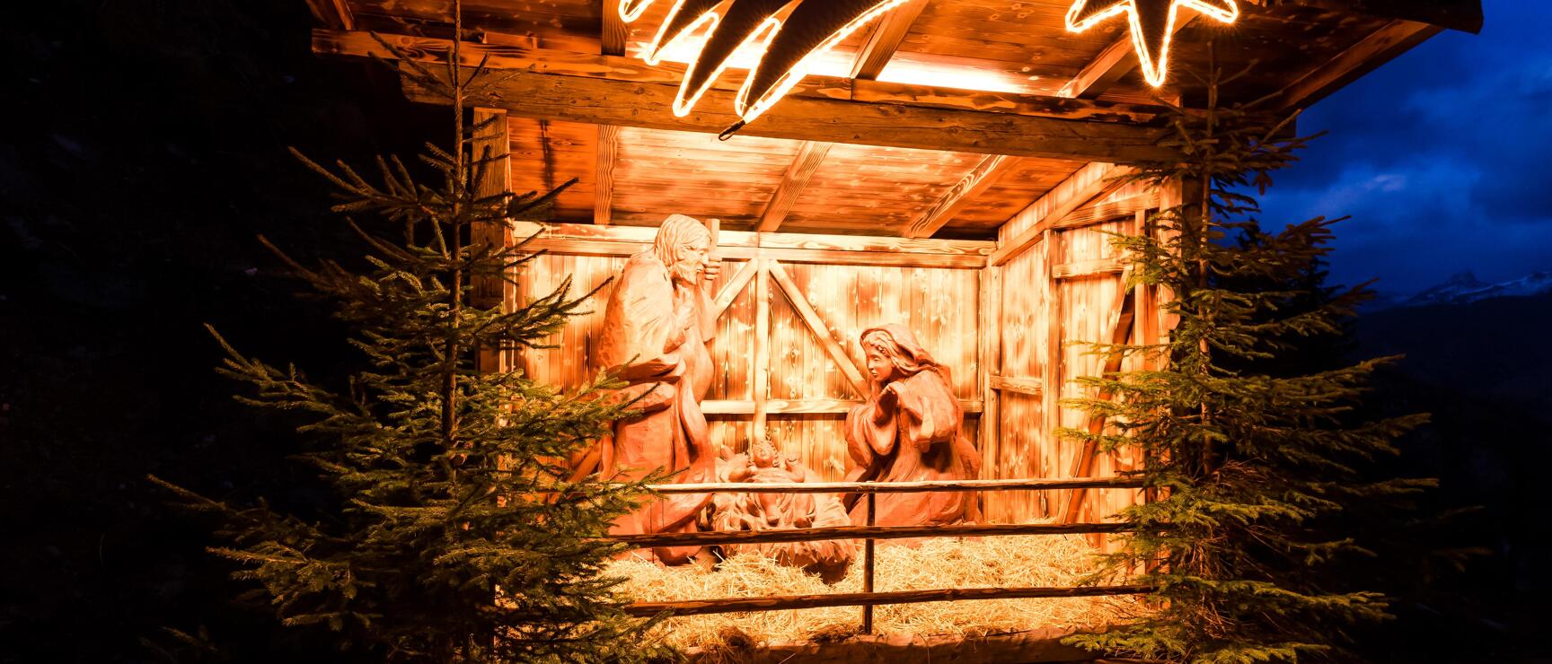 Beleuchtete Holzkrippe mit geschnitzten Figuren und leuchtendem Stern bei Nacht, flankiert von Tannen.