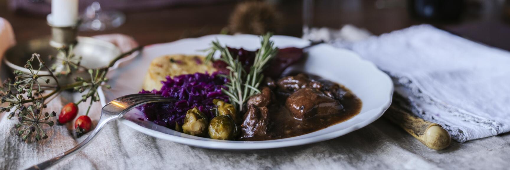 Plate with game ragout, red cabbage and dumplings, red wine and candle on set table.