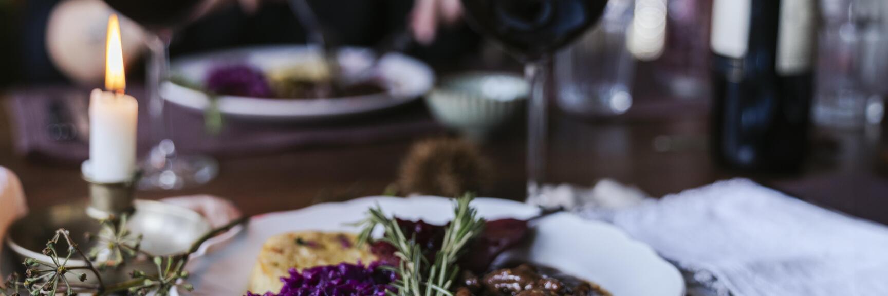 Plate with game ragout, red cabbage and dumplings, red wine and candle on set table.